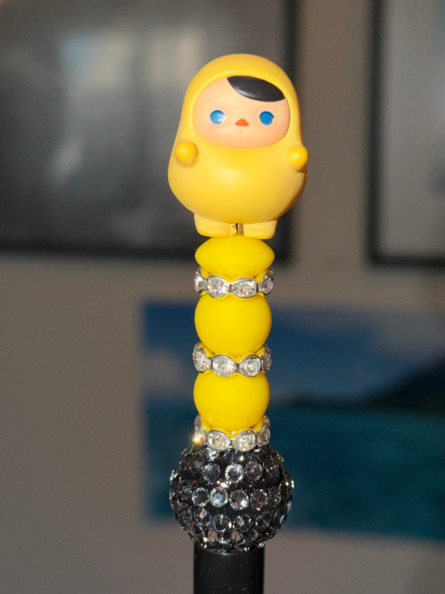 Kawaii beaded pen topped with an authentic Pucky bean yellow hoodie baby figure with black bangs and blue eyes, stacked on bright yellow matte beads with silver rhinestone spacers and a black gunmetal rhinestone disco bead on a black pen barrel