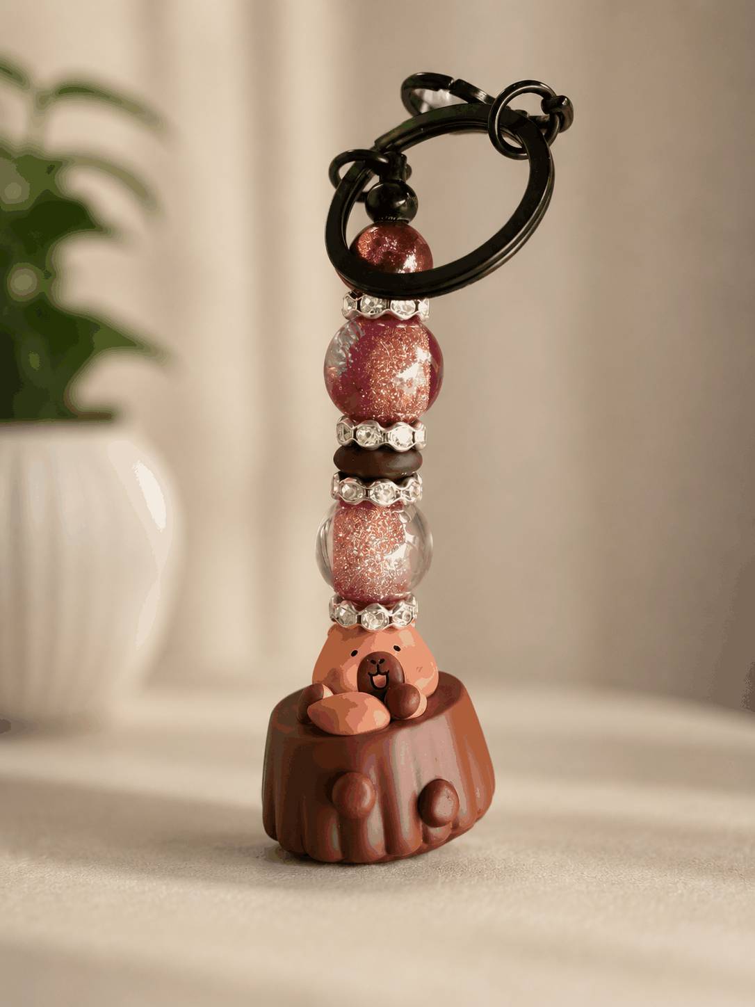 Handmade capybara beaded keychain featuring pink and purple glitter beads, rhinestone spacers, black keyring hardware, and a cute capybara sitting inside a chocolate
