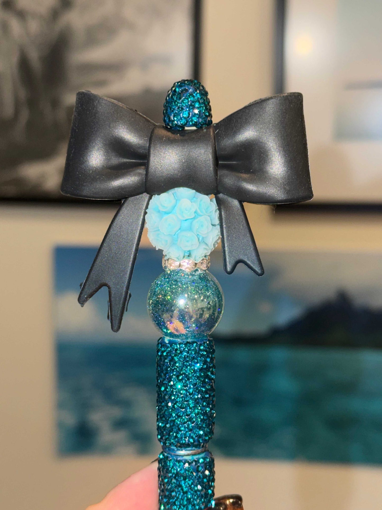 A decorative ballpoint pen held upright. The barrel is fully covered in teal blue rhinestones. Stacked above it are three beads: a glittery iridescent globe, a light blue floral resin ball made of tiny roses, and a small teal crystal-encrusted bead. A large matte black satin bow sits at the top, spread wide like a ribbon bow on a gift.