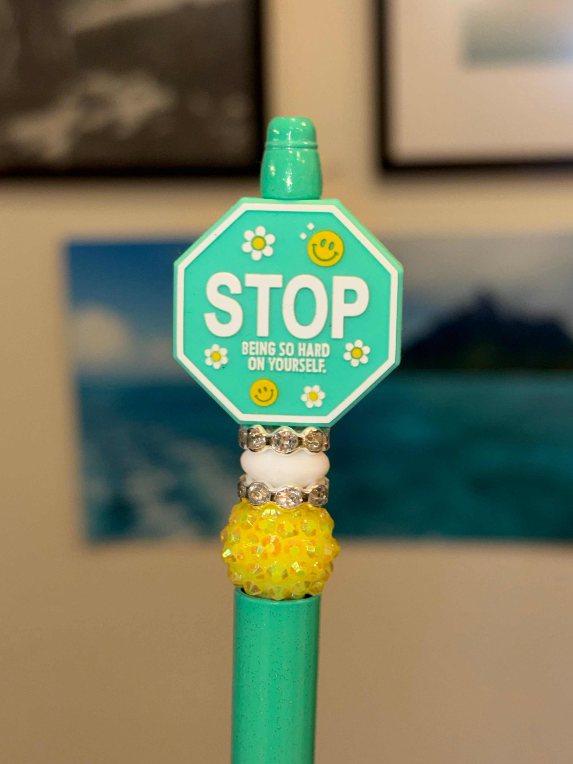 Teal pen with yellow rhinestone bead and white beads with rhinestone spacers and teal stop sign with a white background that says stop being so hard on yourself with white writing and white flowers and smileys 