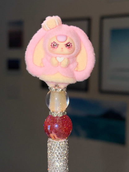 Close-up of a decorative pen topper of a plush toy inspired by a pink and yellow baby 3 with bunny ears and a crown and yellow hair and colorful beads on a full silver bling pen. 