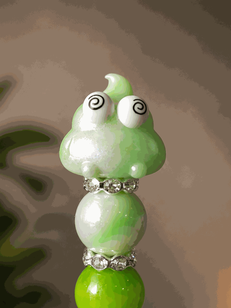 Handmade green beaded pen featuring a cute kawaii poop character with spiral eyes, accented with pearlized green and white beads and sparkling rhinestone spacers, displayed as a fun novelty desk accessory.