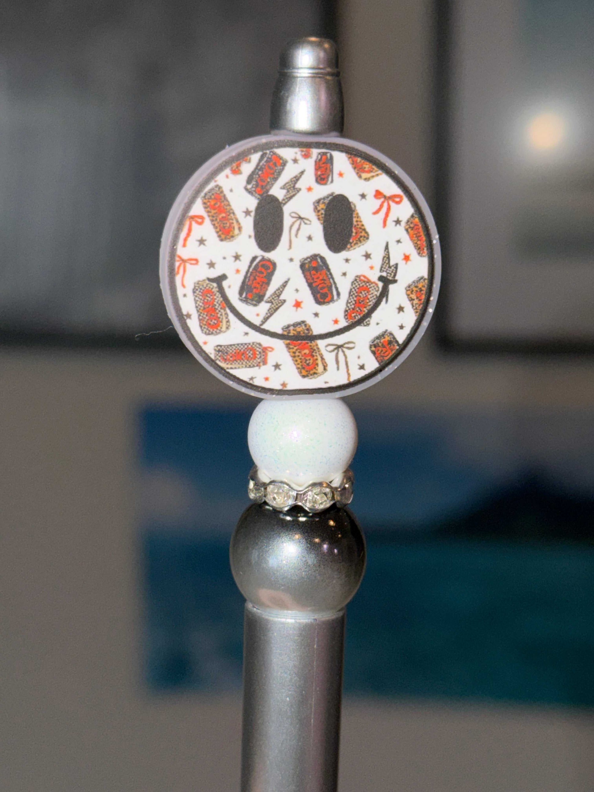 The bead stack includes:
∙ Round acrylic soda smiley face charm topper with diet cokes and coke zeros 
∙ Iridescent glitter white round bead
∙ Silver rhinestone rondelle spacer
∙ Silver metallic round bead
∙ Silver ballpoint pen base
