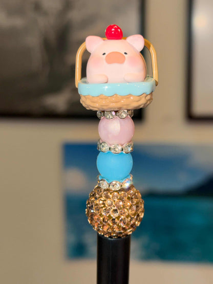 Pink pig 3-D cartoon Lulu pig Bean in a picnic basket with blue towel and strawberry on head on top of pink and blue beads and rhinestones on top of a black pen