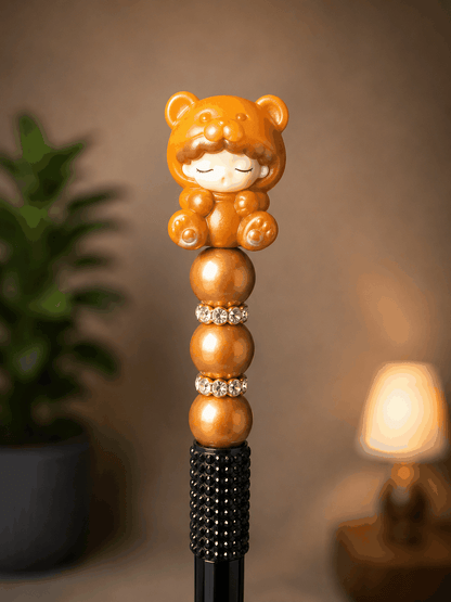Handmade beaded pen featuring an authentic YUMO Gummy Bear Bean collectible topper dressed in a caramel-colored bear costume, accented with metallic bronze beads, rhinestone spacers, and a black half rhinestone textured pen barrel.
