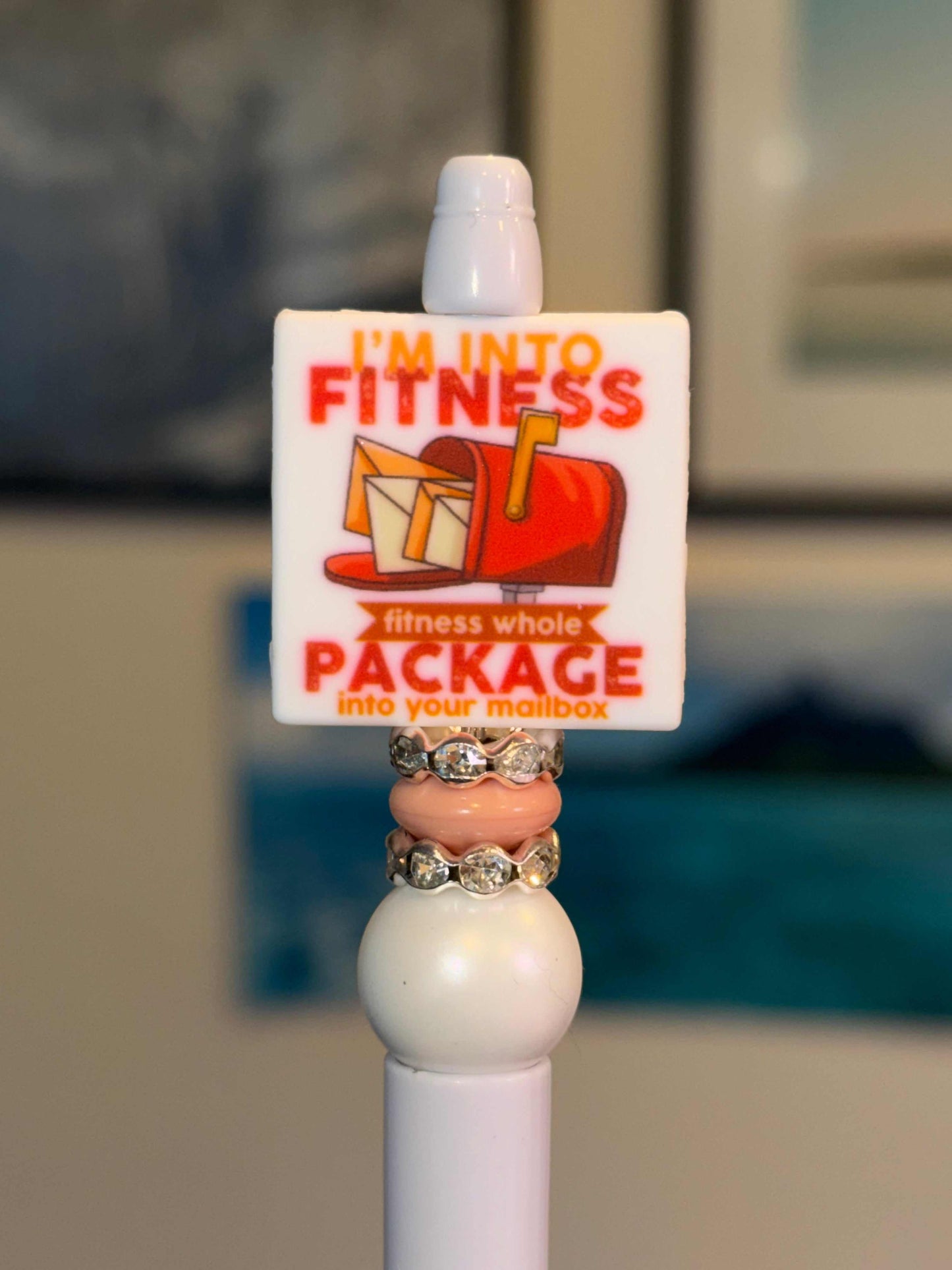 White beaded pen with funny fitness package mailbox quote charm, blush pink donut bead, white bubblegum bead, and clear rhinestone rondelles​​​​​​​​​​​​​​​​  Square silicone focal charm — white background, red & orange “I’m Into Fitness… fitness whole Package into your mailbox” with mailbox graphic