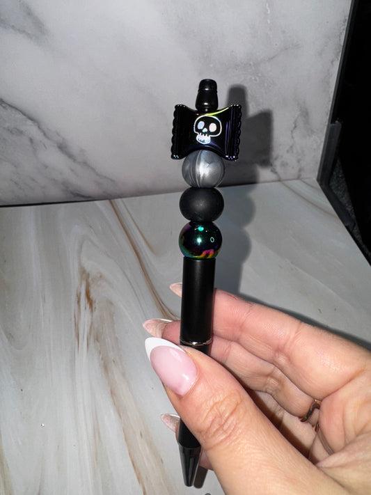 A black beaded pen with black holographic beads with a rainbow hue in certain light. On top sits a Halloween candy wrapper with a skull on it in the same black holographic bead color. 