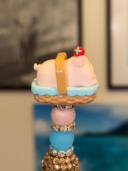 Pink pig 3-D cartoon Lulu pig Bean in a picnic basket with blue towel and strawberry on head on top of pink and blue beads and rhinestones on top of a black pen. Side view 