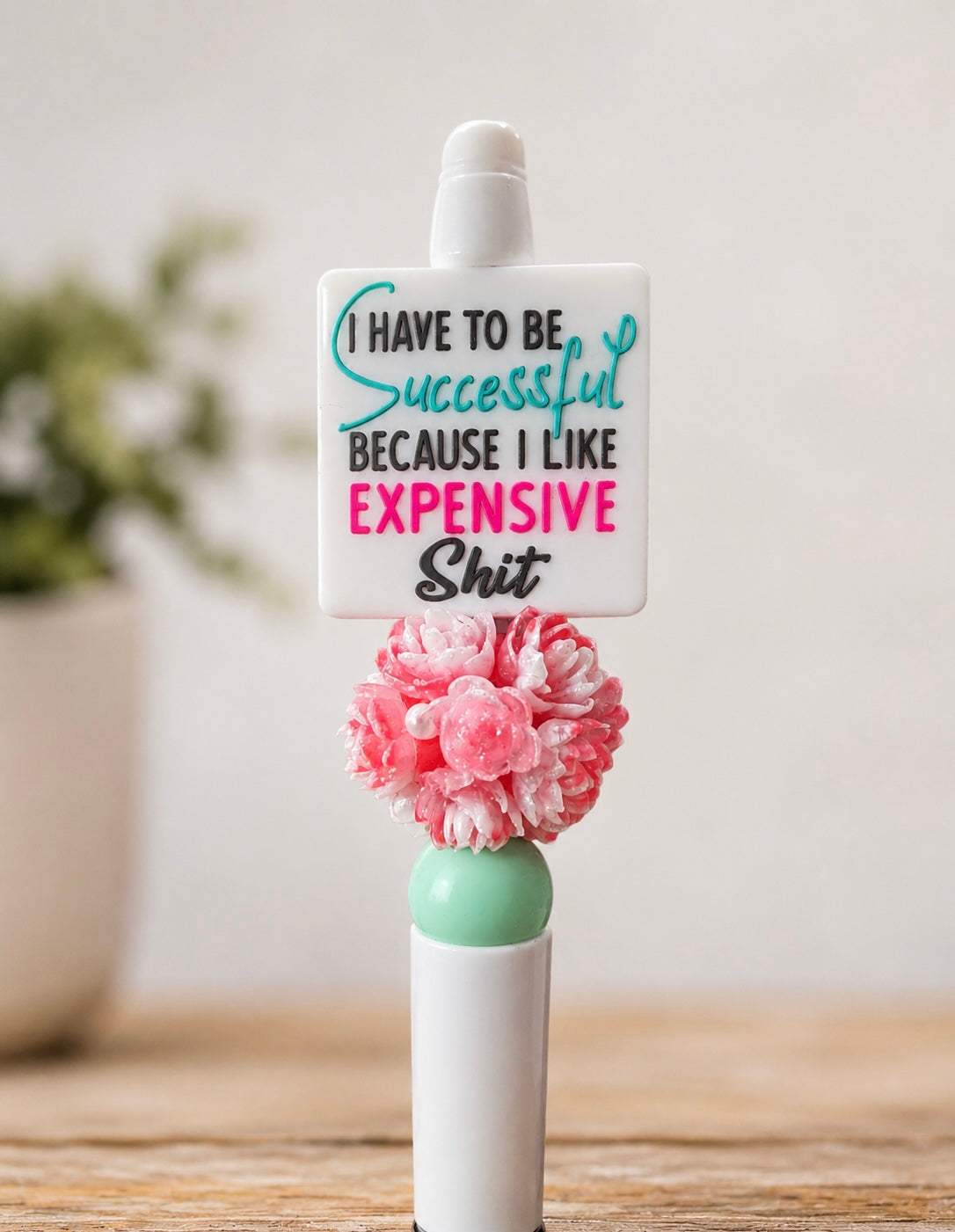 White handmade beaded pen featuring a funny motivational quote topper that reads “I Have to Be Successful Because I Like Expensive Sh*t,” accented with a pink floral bead and mint green bead on a wooden desktop background.