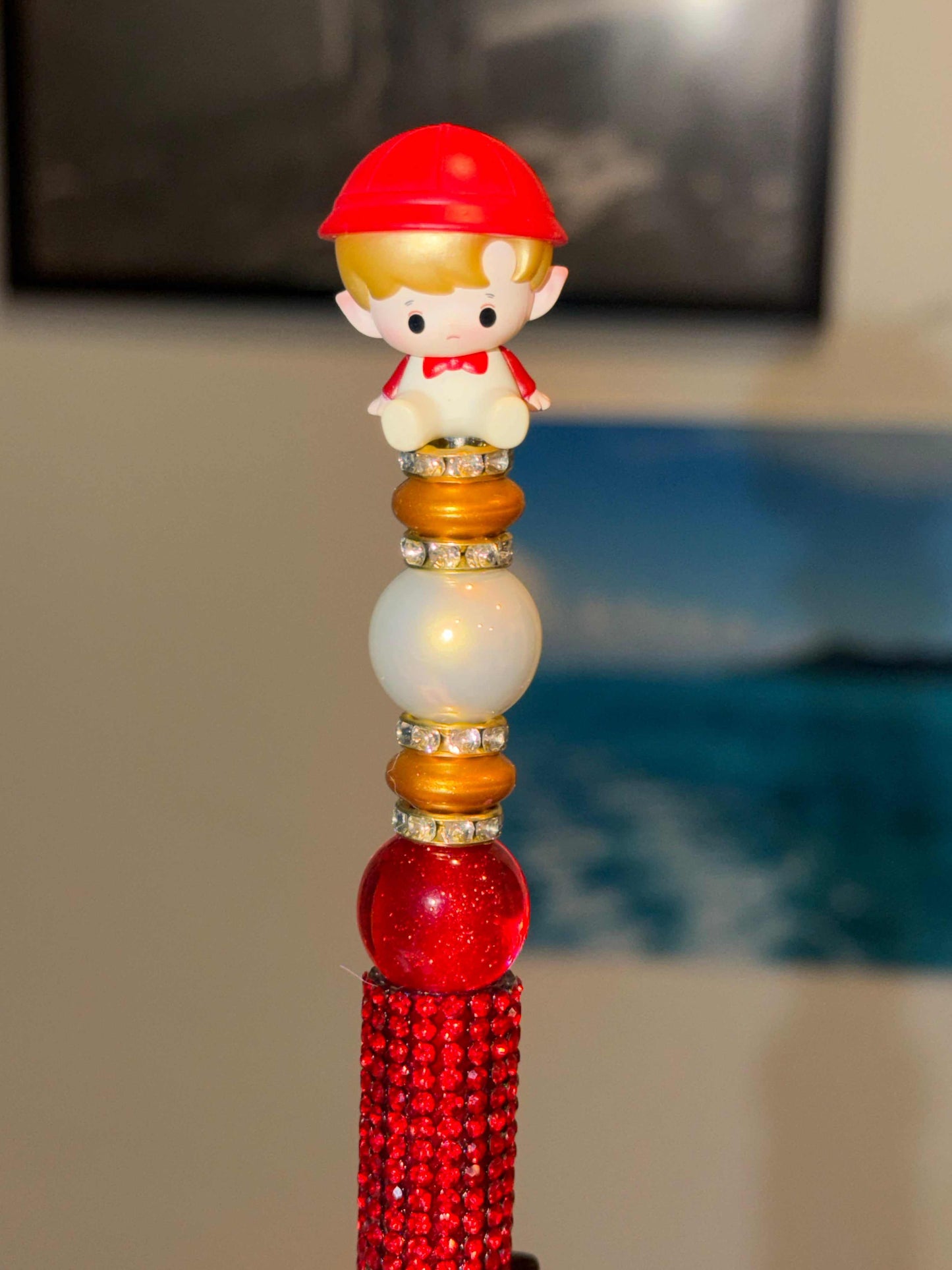 A decorative red rhinestone ballpoint pen photographed upright. From bottom to top: a fully red crystal-encrusted barrel, a deep red glitter globe bead, alternating gold disc beads and clear rhinestone crystal spacers, a large creamy white pearl bead, and more crystal spacers. The pen is topped with a small hacipupu authentic Pop bean collectible figurine wearing a red baseball cap, golden hair, a white rounded body, and a red bow tie, with a soft pouting expression and tiny dangling legs.​​​​​​​​​​​​​​​​