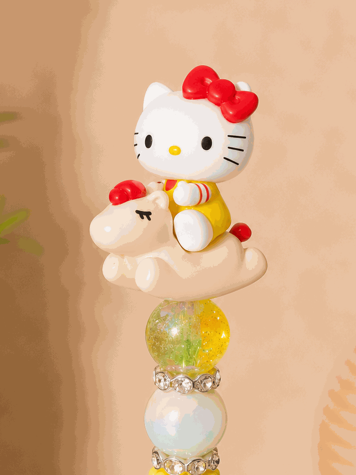 Handmade beaded pen featuring an authentic Hello Kitty Pop Bean figure riding a pony-shaped character topper. The pen includes yellow textured beads, iridescent accent beads, sparkling rhinestone spacers, and a black pen barrel, creating a colorful and playful Sanrio-inspired stationery accessory.