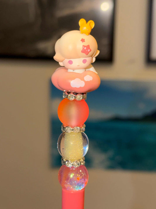 A decorative coral pink ballpoint pen photographed upright. From bottom to top: a coral pink barrel, a clear holographic rainbow glitter globe bead, a crystal rhinestone spacer, a clear yellow shimmer glitter globe bead, another crystal rhinestone spacer, and a large frosted peach-orange matte round bead. The pen is topped with an authentic Dimoo Pop Bean figurine — a cream-toned figure with pink blush cheeks, an orange star detail, and a yellow bunny ear hair clip, nestled into a peach-to-coral bead 