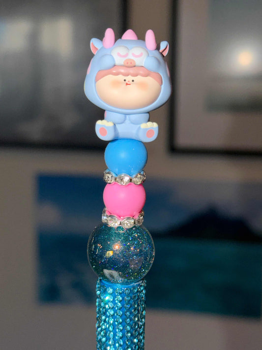 Dino Costume Baby Beaded Pen with blue pink beads and rhinestones, featuring a sleepy baby in blue horned monster onesie topper