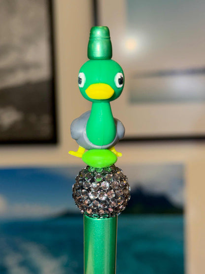 Green and gray 3-D duck cartoon with a yellow beak and yellow feet on green bead and gray rhinestone bead on a green pen. Front profile.