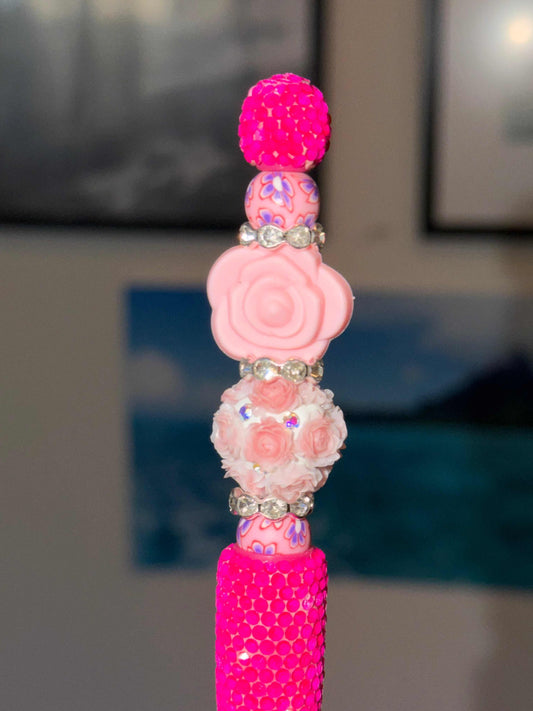 Close-up of a handmade beaded pen with a hot pink rhinestone-wrapped barrel, featuring a bead stack including a hot pink pave disco ball bead, pink floral polymer clay beads, a blush sculpted rose focal bead, a white dimensional floral bouquet bead, and crystal rhinestone rondelle spacers throughout.​​​​​​​​​​​​​​​​