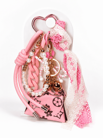 Pink coquette-style bag charm featuring a heart-shaped carabiner, pearl accents, pink lace bow, chunky pink chain links, and a designer-inspired mini purse charm displayed on a white stand against a clean white background.