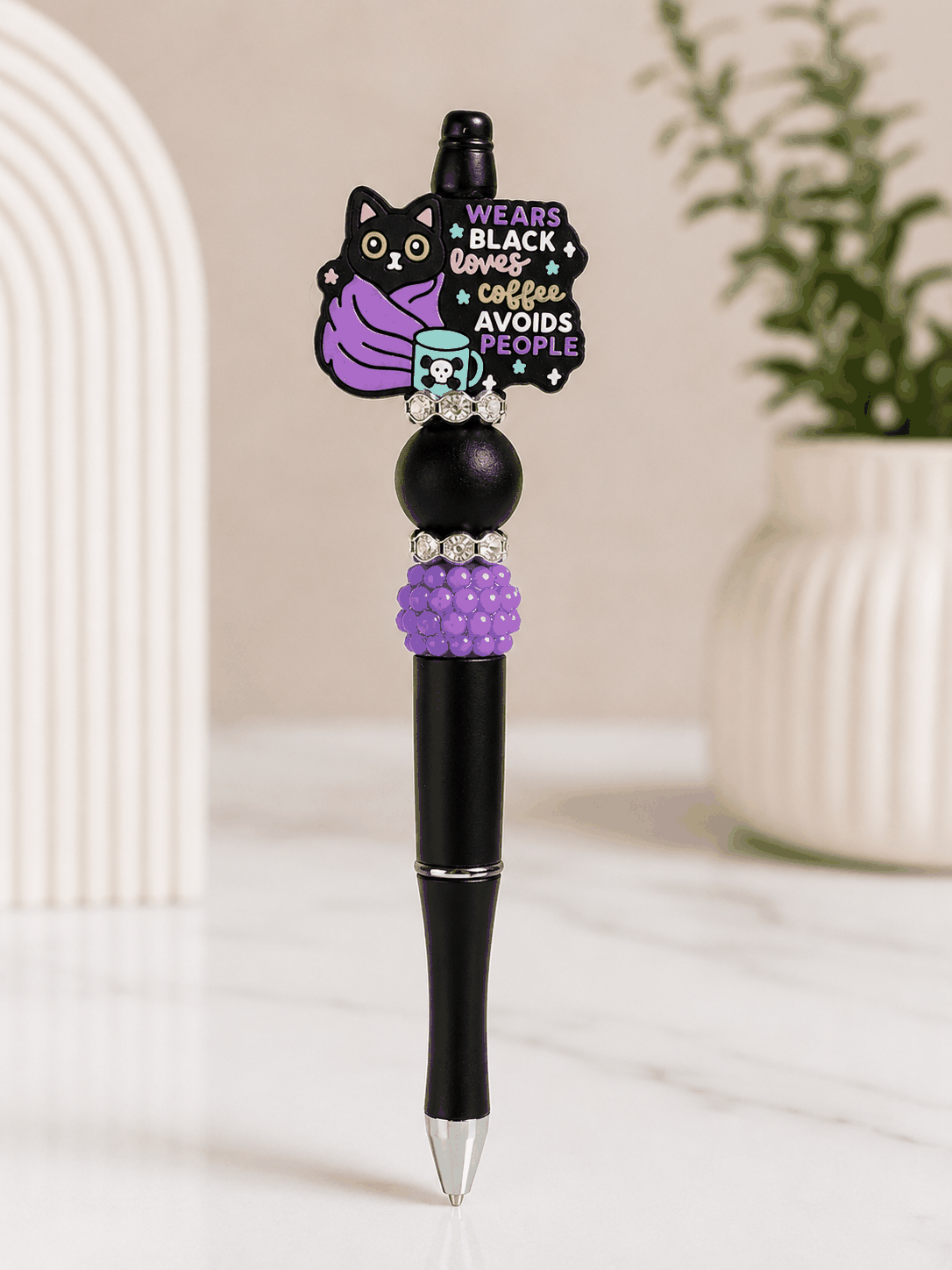 Black beaded pen featuring a black cat wrapped in a purple blanket with a coffee mug and the phrase “Wears Black, Loves Coffee, Avoids People,” accented with purple beads and rhinestone spacers.