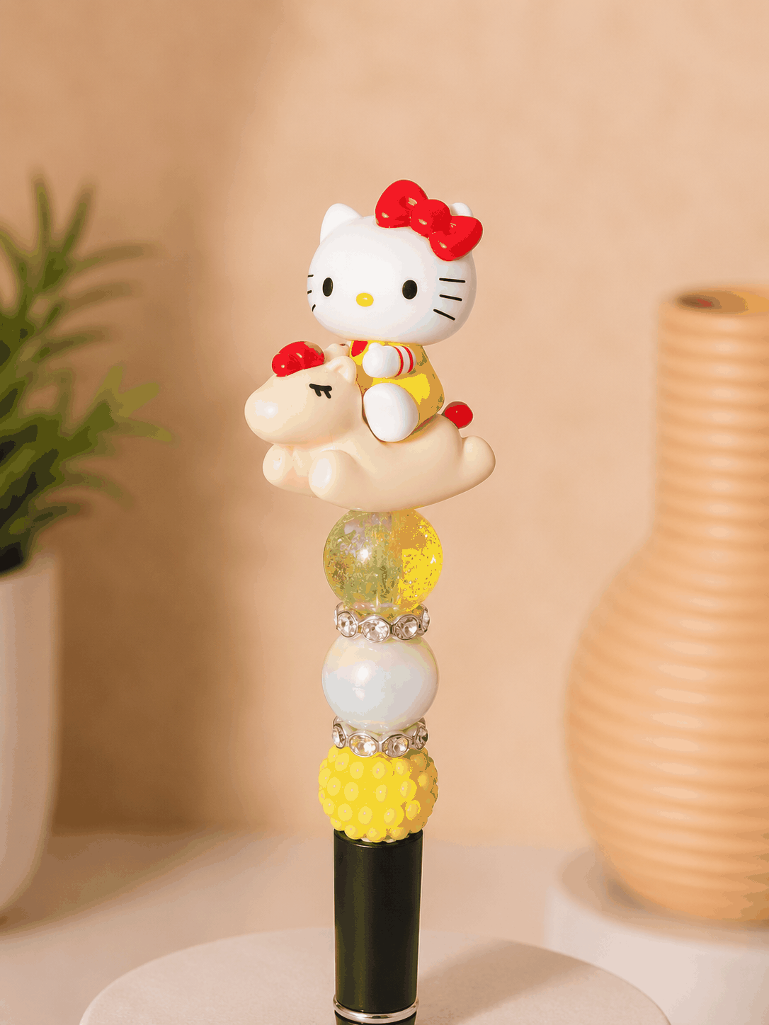Handmade beaded pen featuring an authentic Hello Kitty Pop Bean figure riding a pony-shaped character topper. The pen includes yellow textured beads, iridescent accent beads, sparkling rhinestone spacers, and a black pen barrel, creating a colorful and playful Sanrio-inspired stationery accessory.