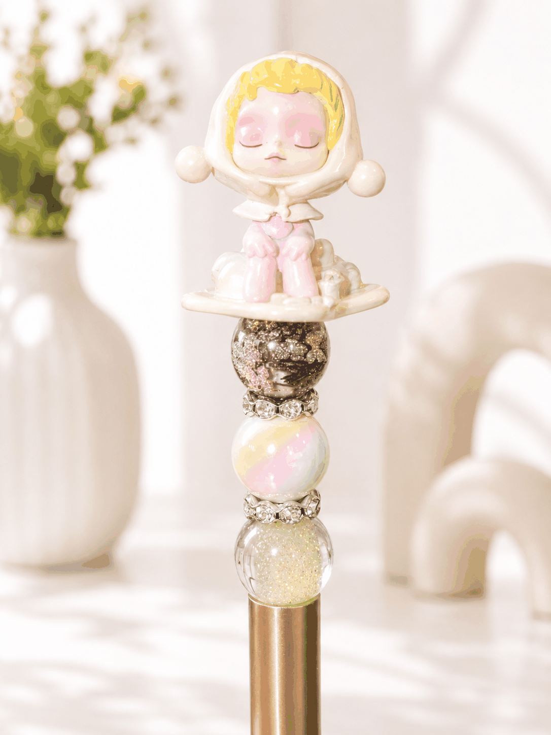 Handmade decorative gold pen featuring a sleeping winter-themed figure that is white and pastel yellow and pastel pink. Inspired by skull panda, with iridescent pastel beads and rhinestone accents.