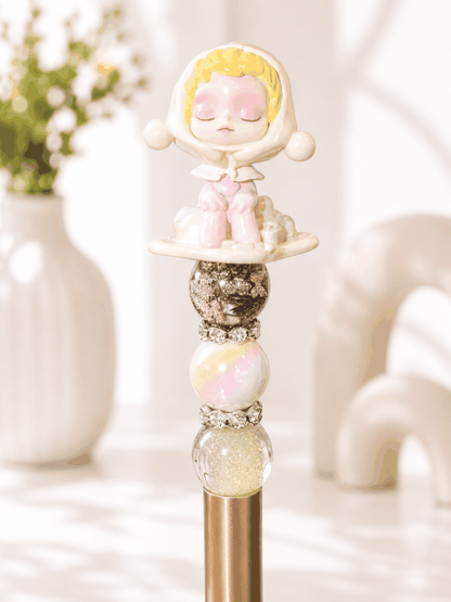 Handmade decorative gold pen featuring a sleeping winter-themed figure that is white and pastel yellow and pastel pink. Inspired by skull panda, with iridescent pastel beads and rhinestone accents.