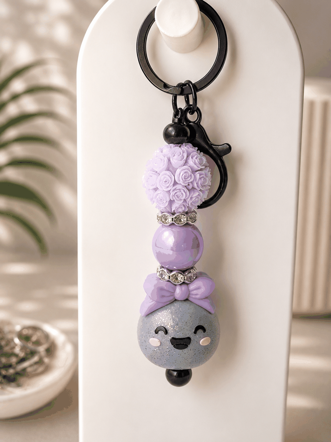 Glitter Dumpling beaded keychain featuring a smiling gray glitter dumpling charm with a lavender bow, lavender floral resin bead, iridescent pearl bead, sparkling rhinestone spacers, and black hardware displayed on a neutral background.