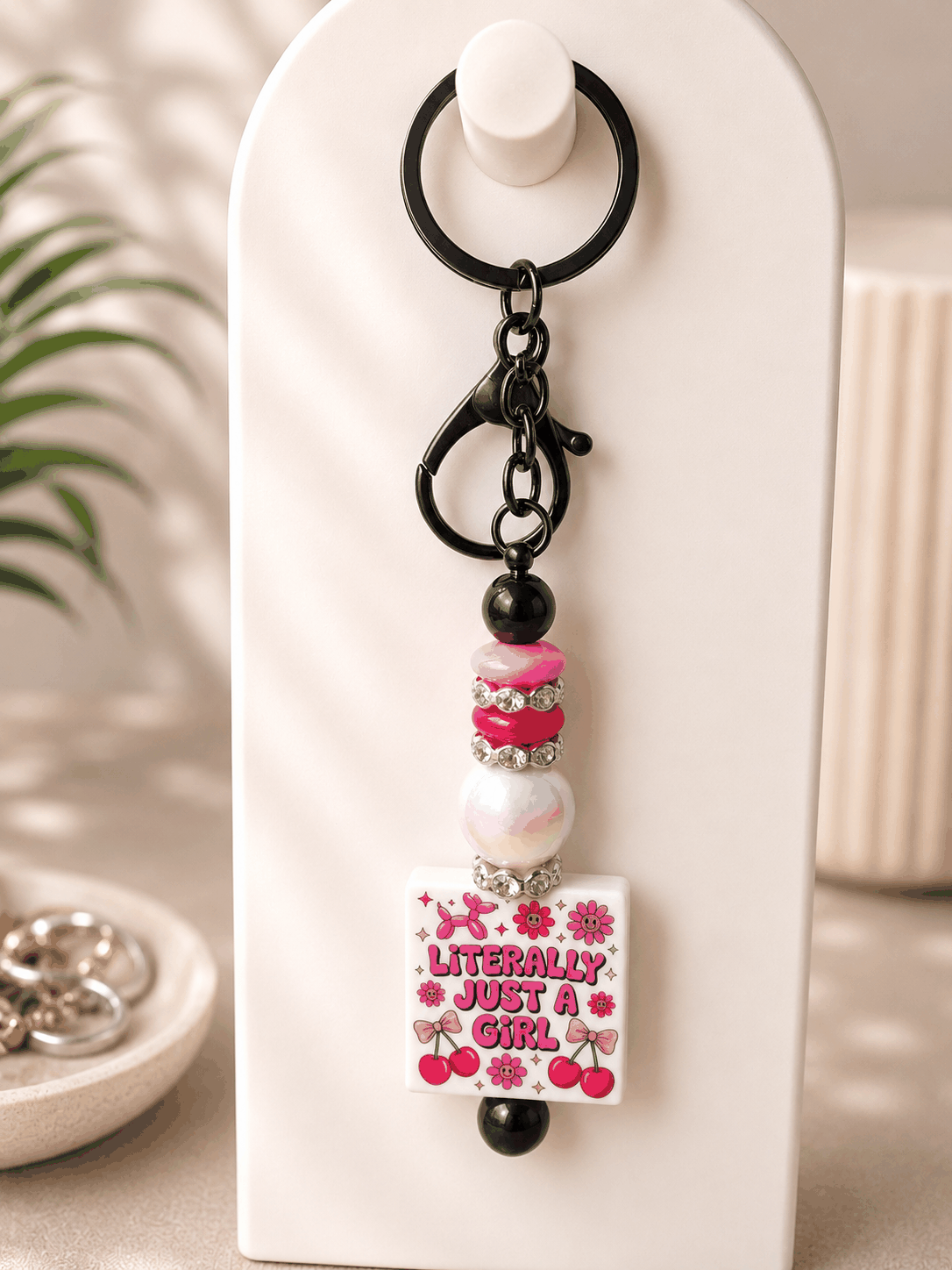 Handmade beaded keychain featuring a “Literally Just a Girl” statement charm decorated with pink flowers and cherries, accented with glossy black beads, pink beads, an iridescent pearl bead, sparkling rhinestone spacers, and black hardware on a neutral background.