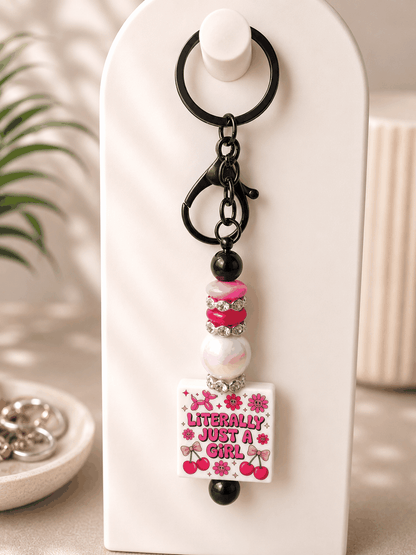 Handmade beaded keychain featuring a “Literally Just a Girl” statement charm decorated with pink flowers and cherries, accented with glossy black beads, pink beads, an iridescent pearl bead, sparkling rhinestone spacers, and black hardware on a neutral background.