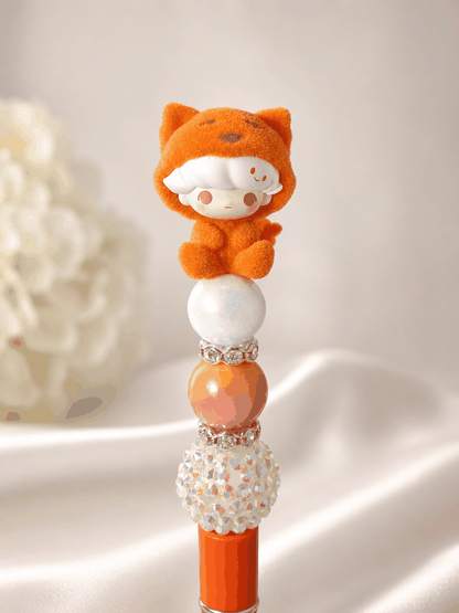 Beaded pen with authentic Pop Mart DIMOO Pop Bean tiger flocked figure topper, iridescent opal bead, peach ombre gradient bead, rhinestone spacer rings, and AB crystal disco ball bead, on an orange pen base, styled against white silk