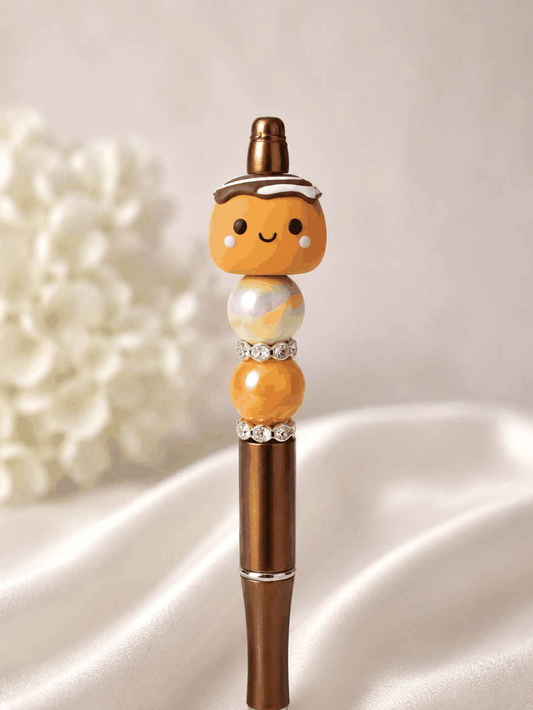 Bronze metallic beaded pen with kawaii cinnamon roll polymer clay focal bead topper with chocolate drizzle and frosting detail, rainbow iridescent swirl bead, warm orange marble bead, and rhinestone spacer rings, styled against white silk