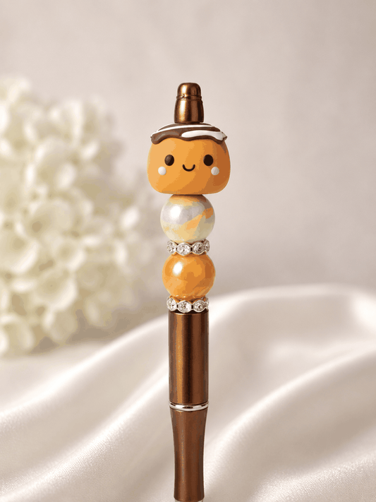 Bronze metallic beaded pen with kawaii cinnamon roll polymer clay focal bead topper with chocolate drizzle and frosting detail, rainbow iridescent swirl bead, warm orange marble bead, and rhinestone spacer rings, styled against white silk