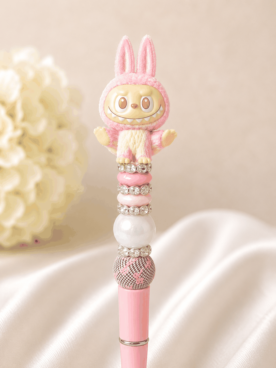 Pink beaded pen with Labubu flocked pink bunny yoga figure topper with orange eyes and wide grin, blush pink rondelle bead, iridescent opal pearl bead, rhinestone spacer rings, and black and white checker bead with pink ribbon print, on a soft pink pen base, styled against white silk