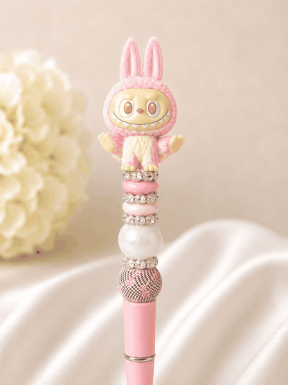 Pink beaded pen with Labubu flocked pink bunny yoga figure topper with orange eyes and wide grin, blush pink rondelle bead, iridescent opal pearl bead, rhinestone spacer rings, and black and white checker bead with pink ribbon print, on a soft pink pen base, styled against white silk