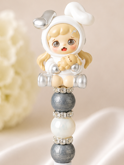 White beaded pen with authentic TNT Dora Bean figure topper in white hoodie onesie with silver robot ears, blonde pigtails, and open-mouthed surprised expression holding a mini teddy bear, slate grey textured matte beads, soft white pearl bead, and rhinestone spacer rings, styled against white silk