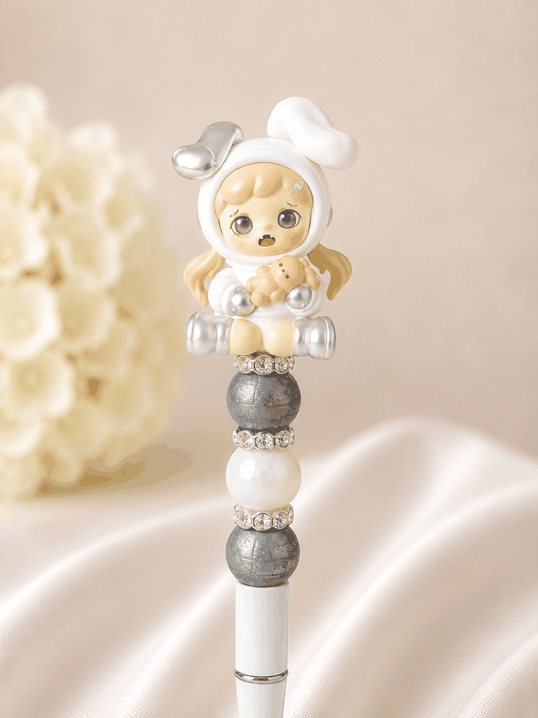 White beaded pen with authentic TNT Dora Bean figure topper in white hoodie onesie with silver robot ears, blonde pigtails, and open-mouthed surprised expression holding a mini teddy bear, slate grey textured matte beads, soft white pearl bead, and rhinestone spacer rings, styled against white silk