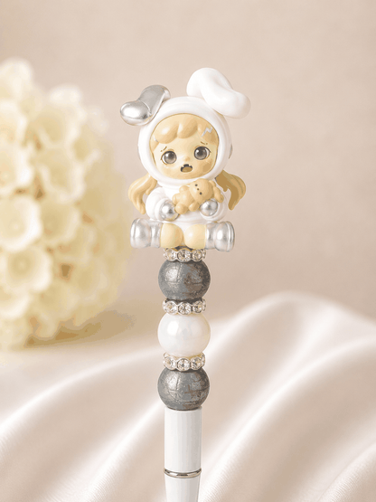 White beaded pen with authentic TNT Dora Bean figure topper in white hoodie onesie with silver robot ears, blonde pigtails, and open-mouthed surprised expression holding a mini teddy bear, slate grey textured matte beads, soft white pearl bead, and rhinestone spacer rings, styled against white silk