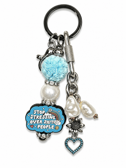 Handmade funny motivational bag charm featuring a blue floral bead, freshwater pearls, sunflower charm, blue crystal heart pendant, and silver hardware on a white background. the funny charm says stop stressing over shitty people. 