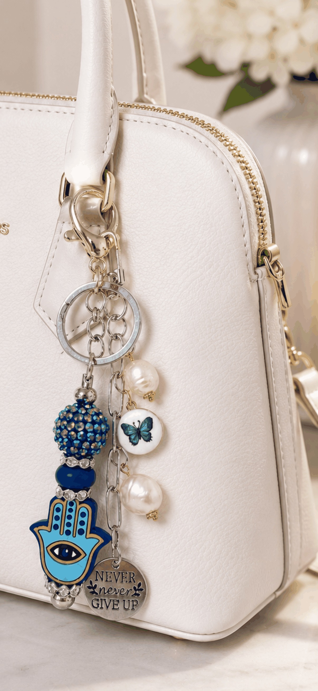 White handbag with decorative keychain featuring a hamsa hand charm, butterfly, and pearls.