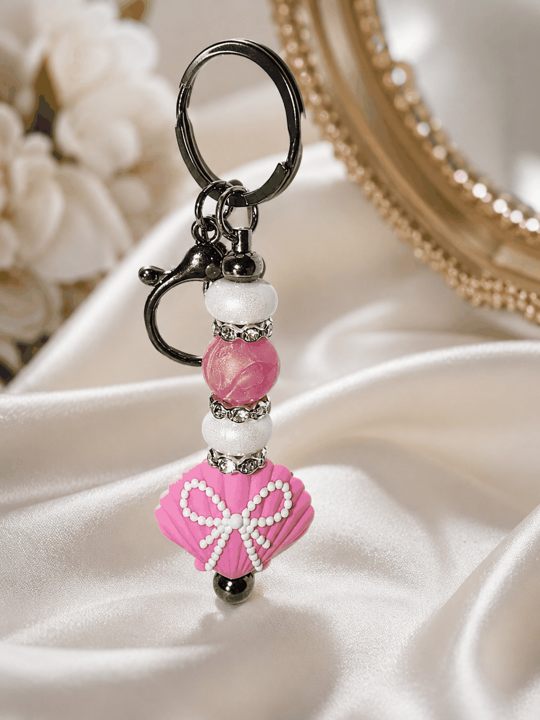 Handmade pink seashell beaded bag charm keychain with pearl-style beads, rhinestone spacers, and black clasp displayed on a luxury satin background.