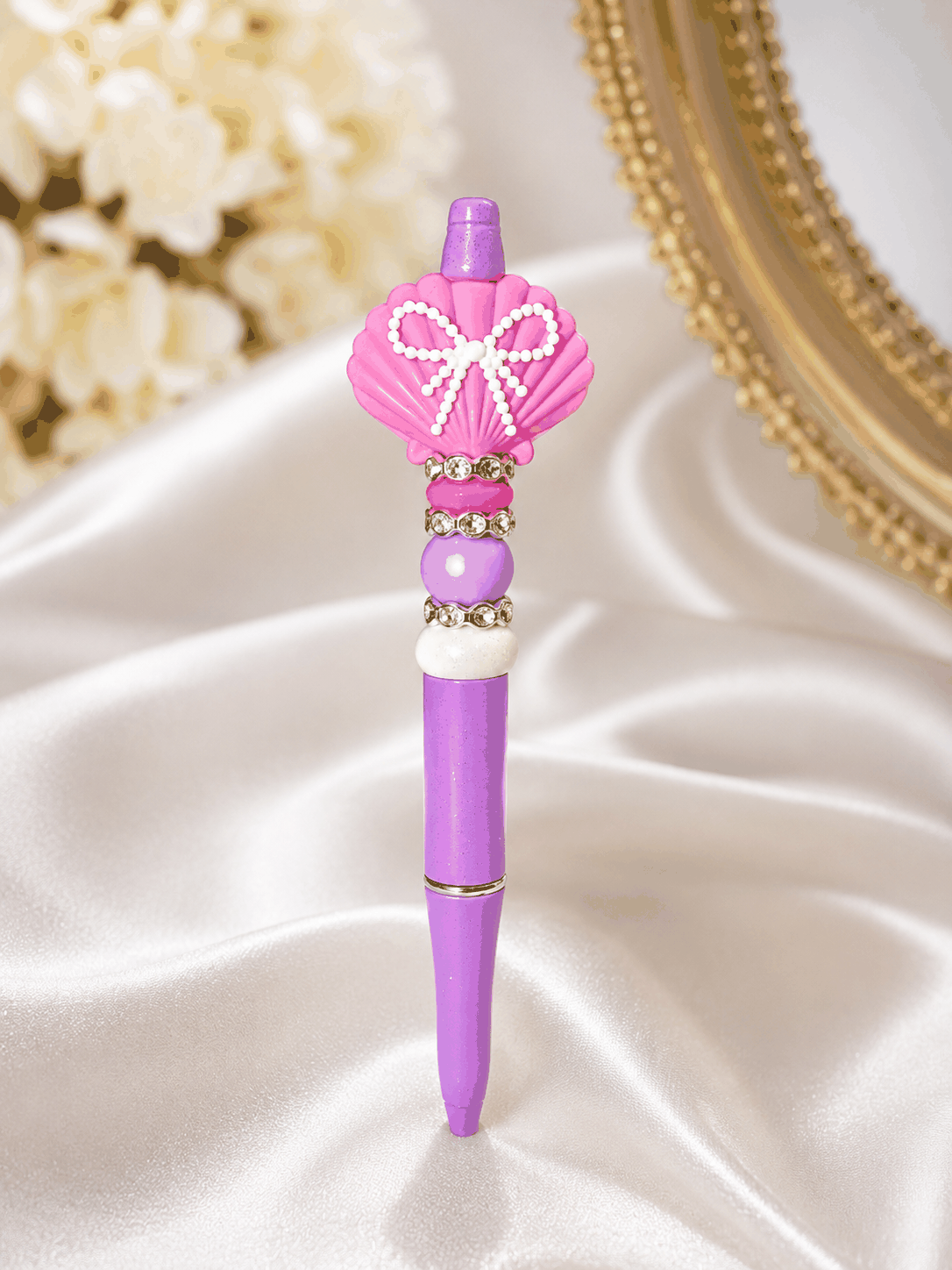Handmade glitter lavender beaded pen featuring a pink seashell focal bead with a white bow design, pastel accent beads, and rhinestone spacers displayed on a luxury satin background.